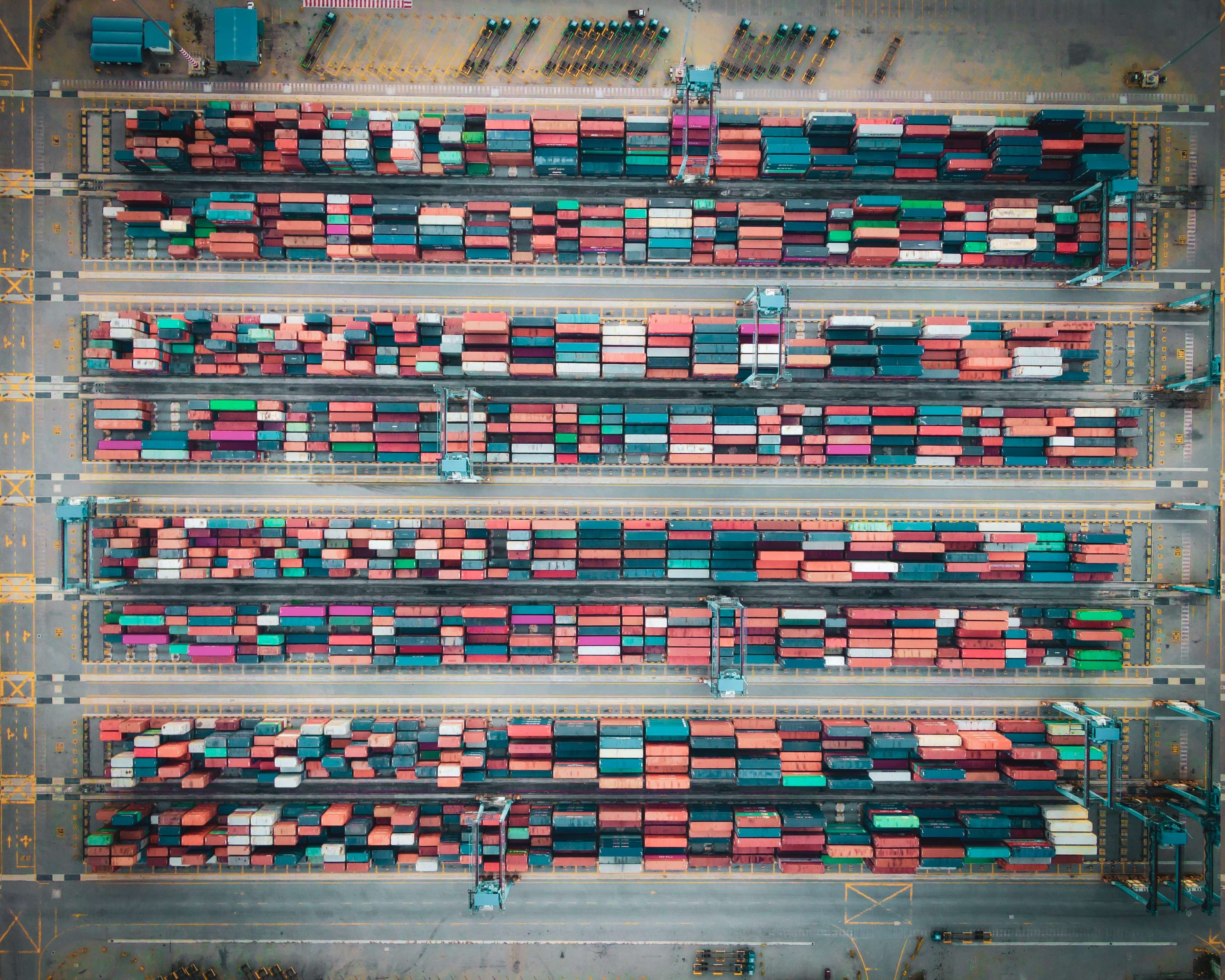 Aerial view of many shipping containers, mostly blue and red, stacked neatly in a large yard. The image represents logistics and trade.